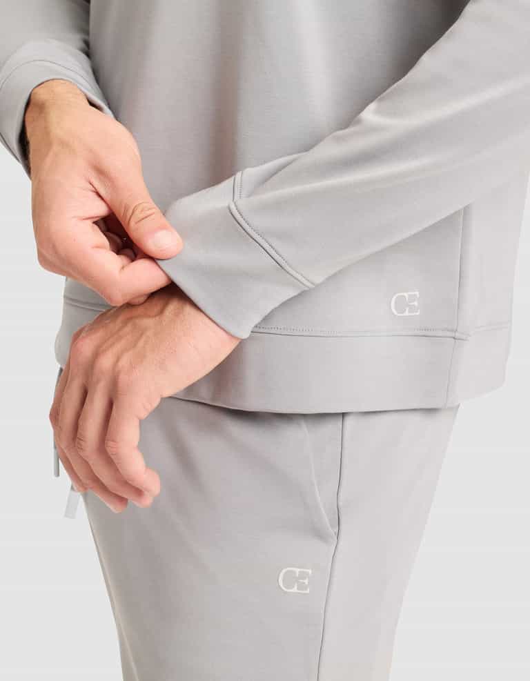 A person wearing Cozy Earth’s Men’s Brushed Bamboo Pullover Crew in light gray adjusts the sleeve. The sweatshirt features a small "CE" logo. Only their torso and hands are visible against a plain background. |Color:Stone
