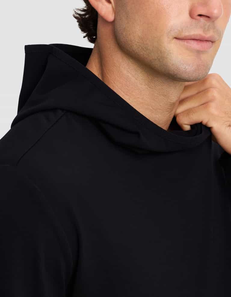 A close-up shows a person wearing the Cozy Earth Men's Brushed Bamboo Recovery Hoodie in black, gently holding the hood's edge near their neck, with their face partially visible and a slight smile.|Color:Black