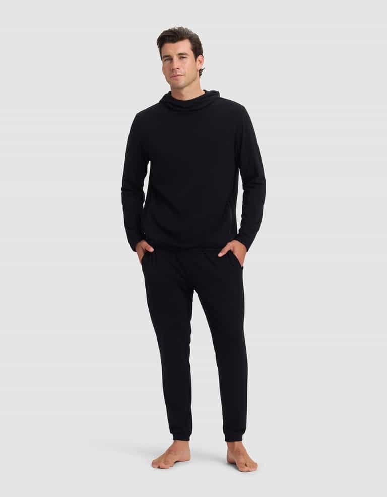 A man stands barefoot against a plain background, wearing the Cozy Earth Men's Brushed Bamboo Recovery Hoodie & Jogger Set in black. His hands are in his pockets and he gazes forward with a neutral expression.|Color:Black