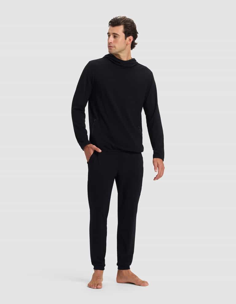 A man stands barefoot against a plain background, wearing the Cozy Earth Men's Brushed Bamboo Recovery Hoodie & Jogger Set in black, featuring a loose-neck hoodie and jogger pants. He looks slightly to the side with a relaxed posture.|Color:Black