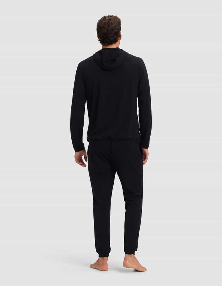 A man stands barefoot facing away, wearing the Cozy Earth Men's Brushed Bamboo Recovery Hoodie & Jogger Set in black, against a plain light gray background.|Color:Black