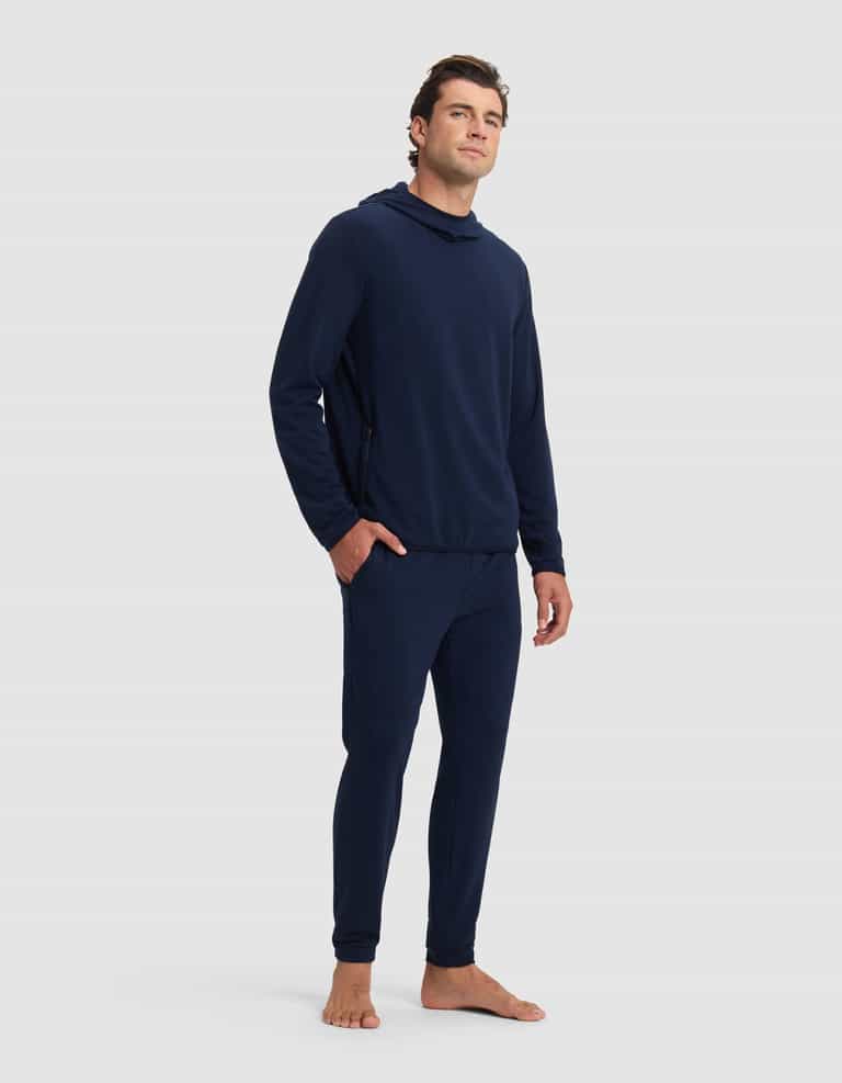 A man with short brown hair stands barefoot against a plain background, wearing the Cozy Earth Men's Brushed Bamboo Recovery Hoodie & Jogger Set in dark blue. He gazes slightly upward with a neutral expression.|Color:Navy