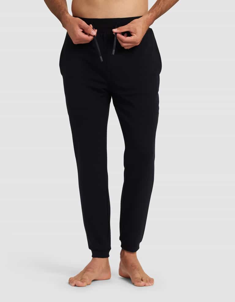 A person stands barefoot on a white background, wearing Cozy Earth's Men's Brushed Bamboo Recovery Jogger in black and adjusting the waistband drawstring. Their upper body is mostly out of frame.|Color:Black