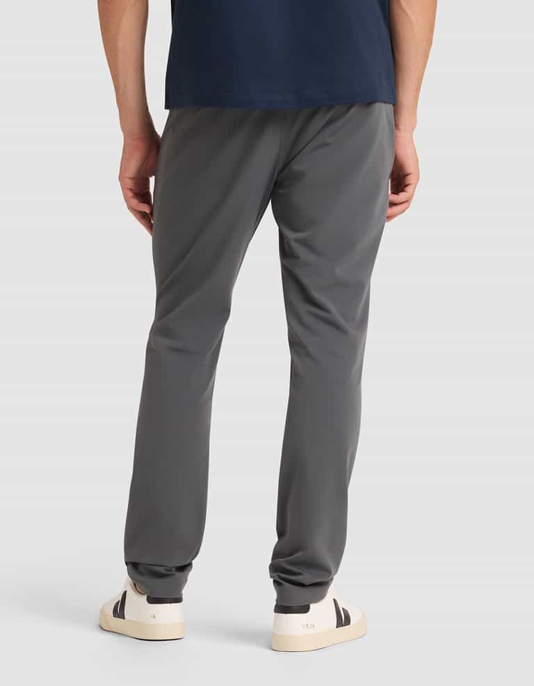 A person stands facing away, wearing Cozy Earth's Men's Everywhere Pant in dark gray, a navy blue shirt, and white sneakers with black heel accents against a plain light background. |Color:Coal