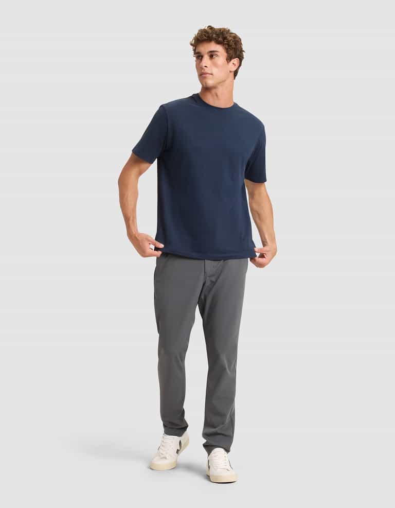A man stands against a light gray background, wearing Cozy Earth's Men's Everywhere Pant in gray, paired with a plain navy blue T-shirt and white sneakers. He looks slightly to the side with a relaxed posture, one hand in his pocket. |Color:Coal
