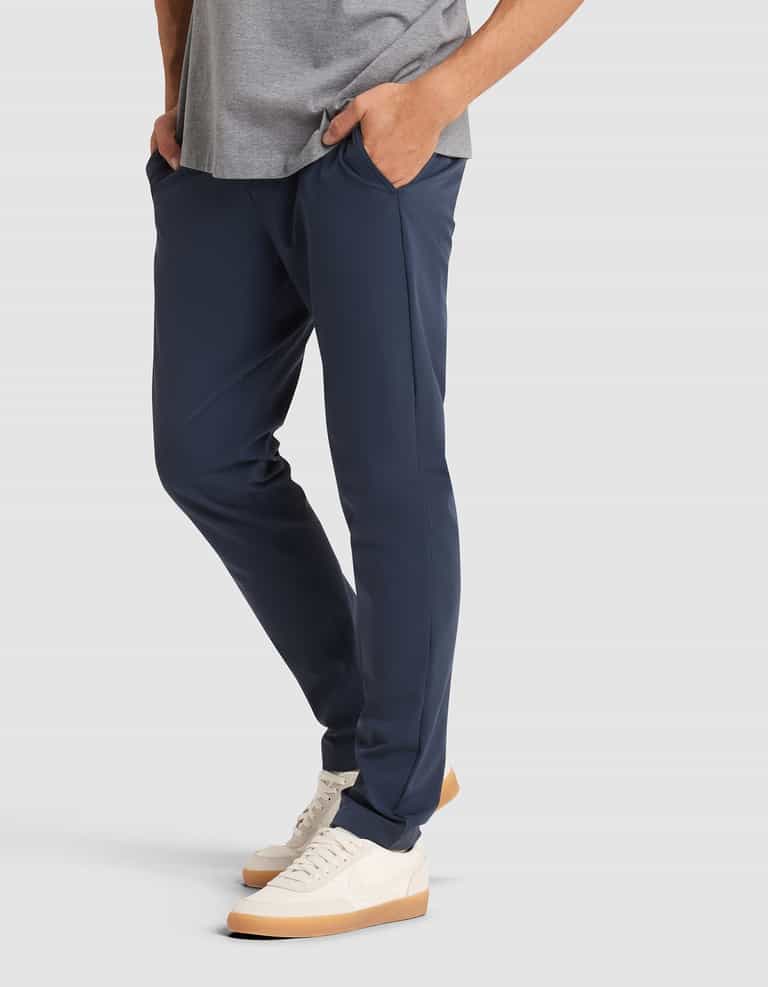A person wearing Cozy Earth's Men's Everywhere Pant in navy, a gray tee, and white sneakers with gum soles stands hands-in-pockets against a light gray background. Only the lower body is shown. |Color:Eclipse