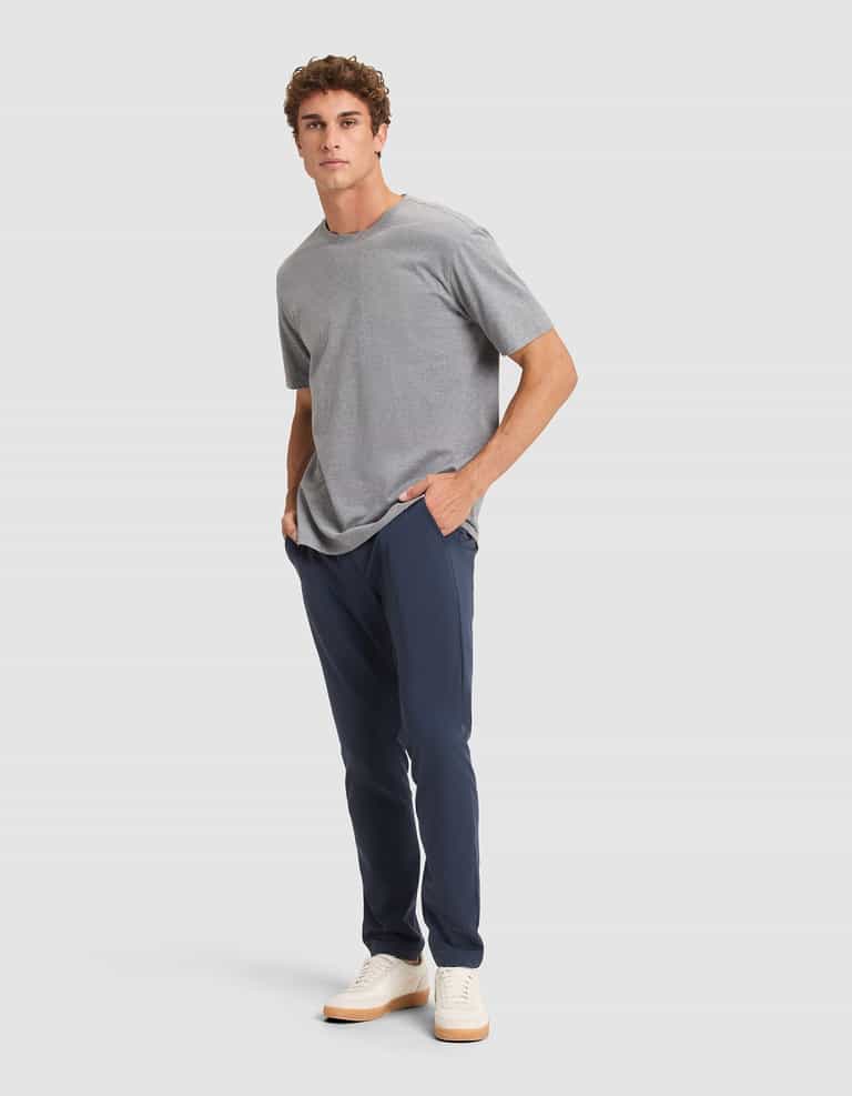A man stands against a light gray background wearing Cozy Earth’s Men's Everywhere Pant in navy, a gray t-shirt, and white sneakers. He has short curly hair, hands in pockets, and looks slightly to the side. |Color:Eclipse