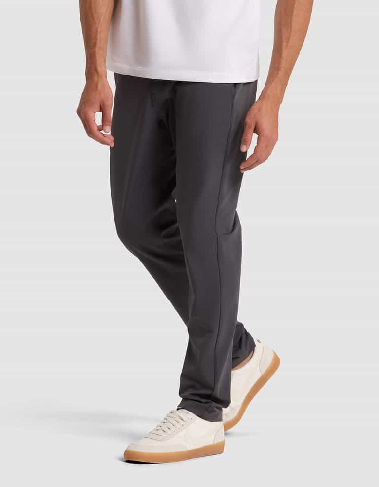 A person wearing Cozy Earth's Men's Everywhere Pant in dark gray, paired with a white shirt and white sneakers with tan soles, stands against a light gray background. Only the lower half of the body is shown. |Color:Graphite