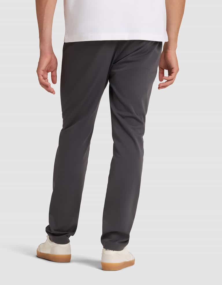 A person is seen from behind, standing against a light background, wearing Cozy Earth's Men's Everywhere Pant in dark gray, paired with a white shirt and white sneakers. |Color:Graphite