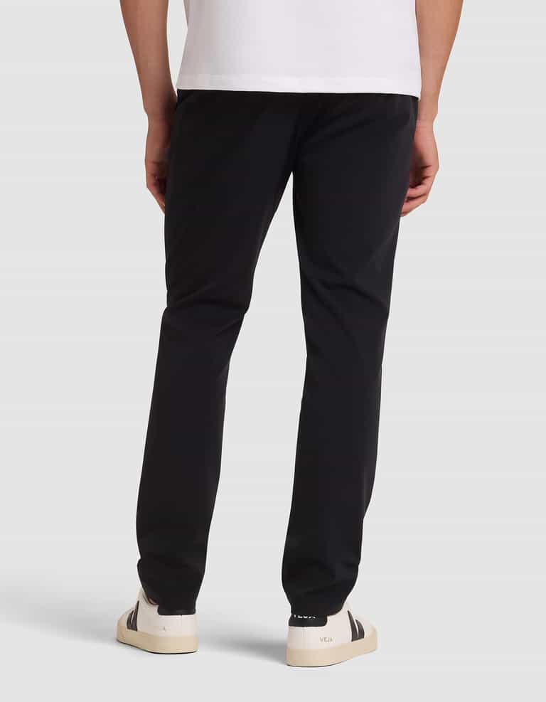 A person stands facing away against a plain light background, wearing Cozy Earth's Men's Everywhere Pant in black, a white t-shirt, and white sneakers with black detailing. |Color:Jet Black