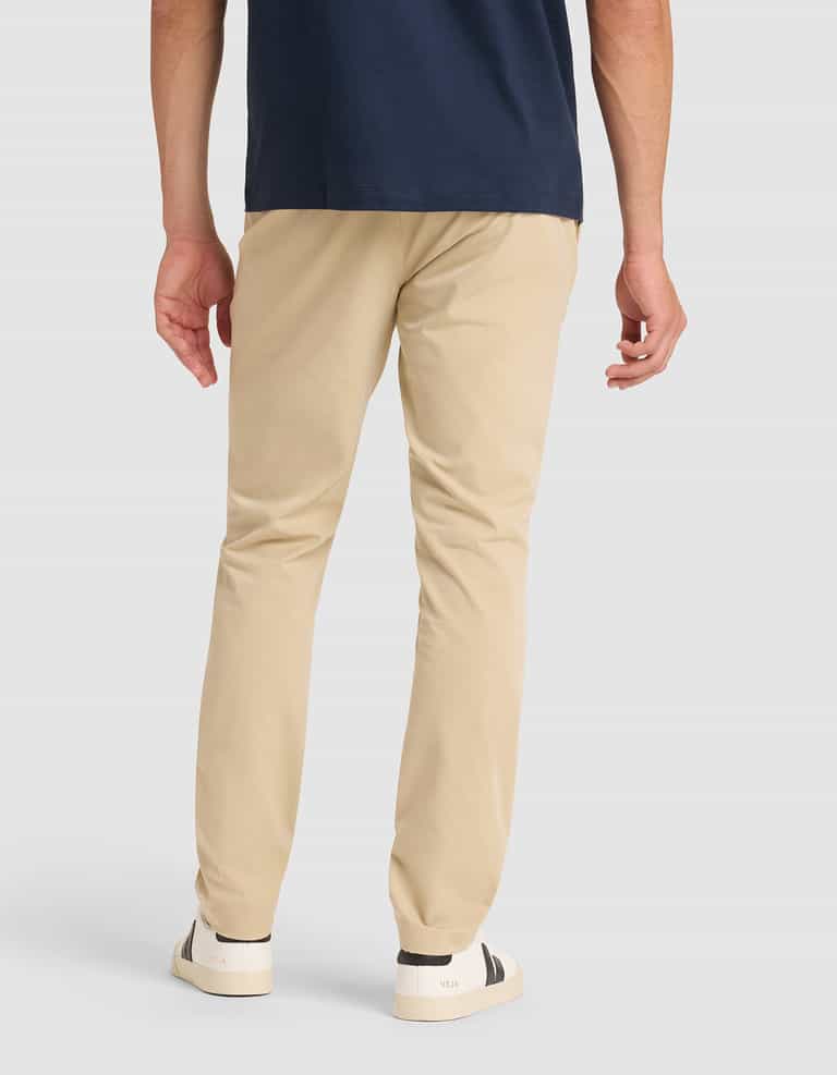 A person stands with their back to the camera, wearing Cozy Earth's Men's Everywhere Pant in beige, a navy shirt, and white sneakers with black accents against a plain light background. |Color:Khaki