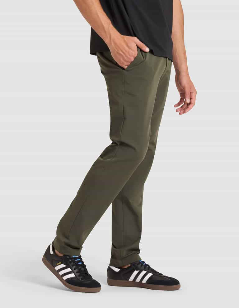A person wearing Cozy Earth's Men's Everywhere Pant in olive green, a black shirt, and black Adidas sneakers with white stripes stands with one hand in their pocket against a plain white background. |Color:Olive