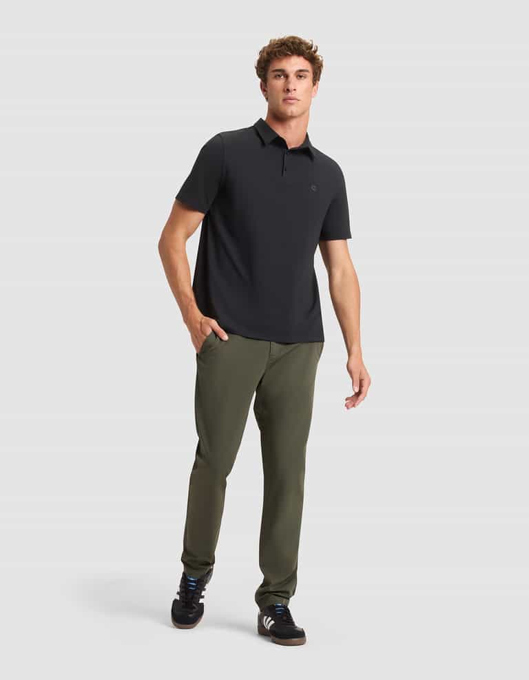 A man stands against a plain light background wearing Cozy Earth's Men's Everywhere Pant in olive green, paired with a black polo shirt and black sneakers. One hand is in his pocket and he has a neutral expression. |Color:Olive