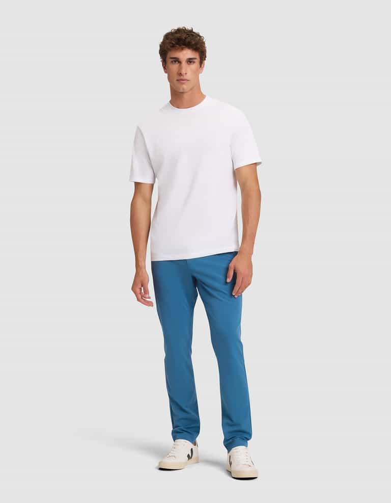 A man stands against a light gray background, wearing Cozy Earth's Men's Everywhere Pant in blue, a plain white short-sleeve t-shirt, and white sneakers. He faces forward with a neutral expression and relaxed posture. |Color:Wave