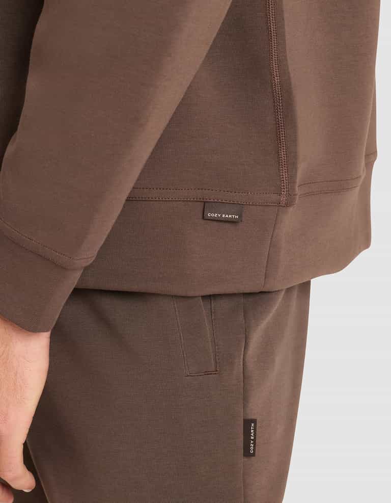 A close-up of a person wearing the Cozy Earth Men's StretchTech Crewneck and matching pants, both featuring small black "COZY EARTH" tags. The person's hand rests at their side. |Color:Cacao