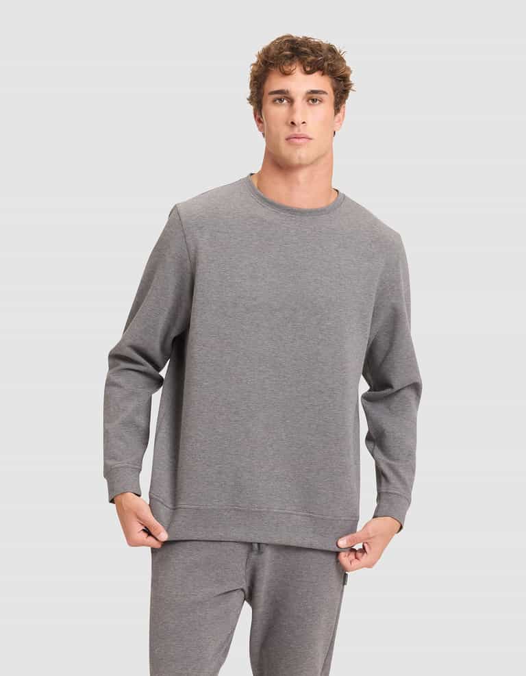 A young man in a Cozy Earth Men's StretchTech Crewneck and matching sweatpants stands before a plain light gray background, looking at the camera while holding the hem of his sweatshirt. |Color:Coal Heather