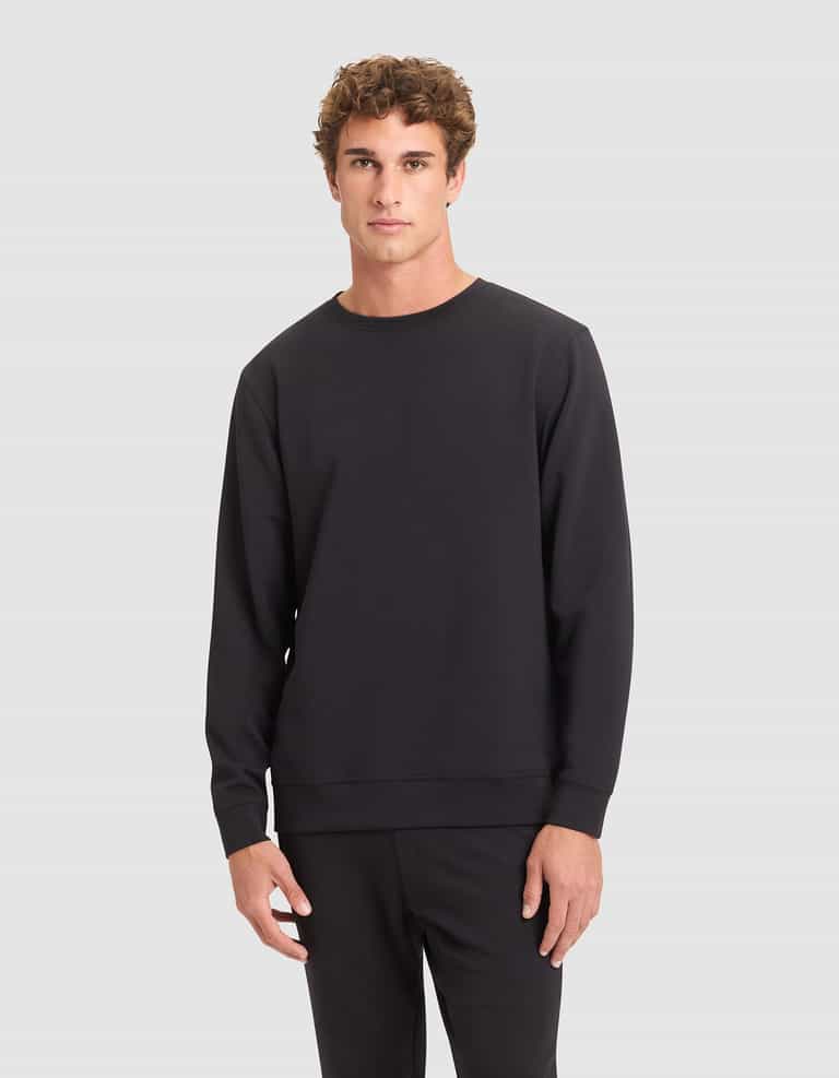 A young man with curly brown hair wears the Cozy Earth Men's StretchTech Crewneck in black with matching black pants, standing against a light gray background. |Color:Jet Black