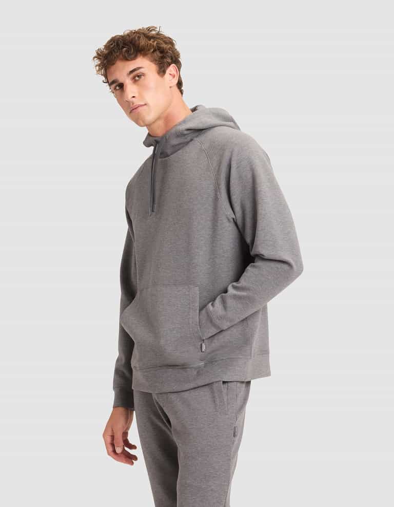 A young man models the Cozy Earth Men's StretchTech Hoodie in light grey, featuring a front pocket and relaxed fit sweatpants, standing against a plain backdrop and gazing slightly to the side. |Color:Coal Heather