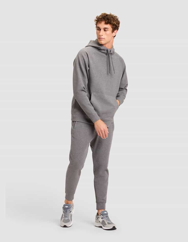 A man stands against a plain background wearing the Cozy Earth Men's StretchTech Hoodie and matching joggers, paired with gray and white sneakers. He has one hand in his pocket and looks to the side. |Color:Coal Heather