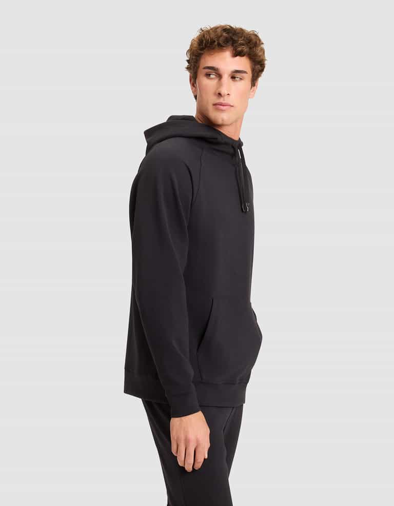 A young man wears the Cozy Earth Men's StretchTech Hoodie in black and matching pants, standing against a plain light gray background while looking to his left with his right hand at his side. |Color:Jet Black