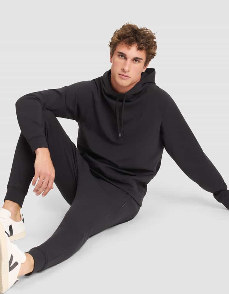 A young man with curly hair wears the Cozy Earth Men's StretchTech Hoodie and matching joggers, paired with white sneakers. He sits on the floor against a light gray background, looking at the camera with one knee up and an arm resting on his leg. |Color:Jet Black