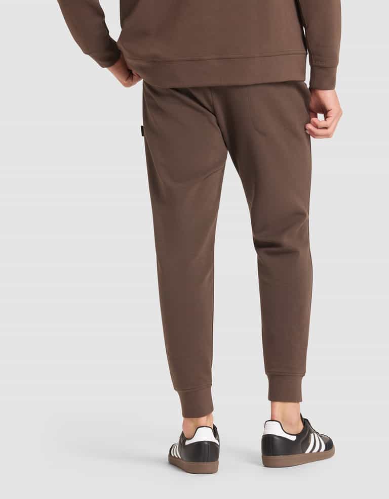 A person wearing Cozy Earth Men's StretchTech Jogger pants and a matching brown sweatshirt, paired with black Adidas sneakers with white stripes, is shown from the back against a plain light background. |Color:Cacao