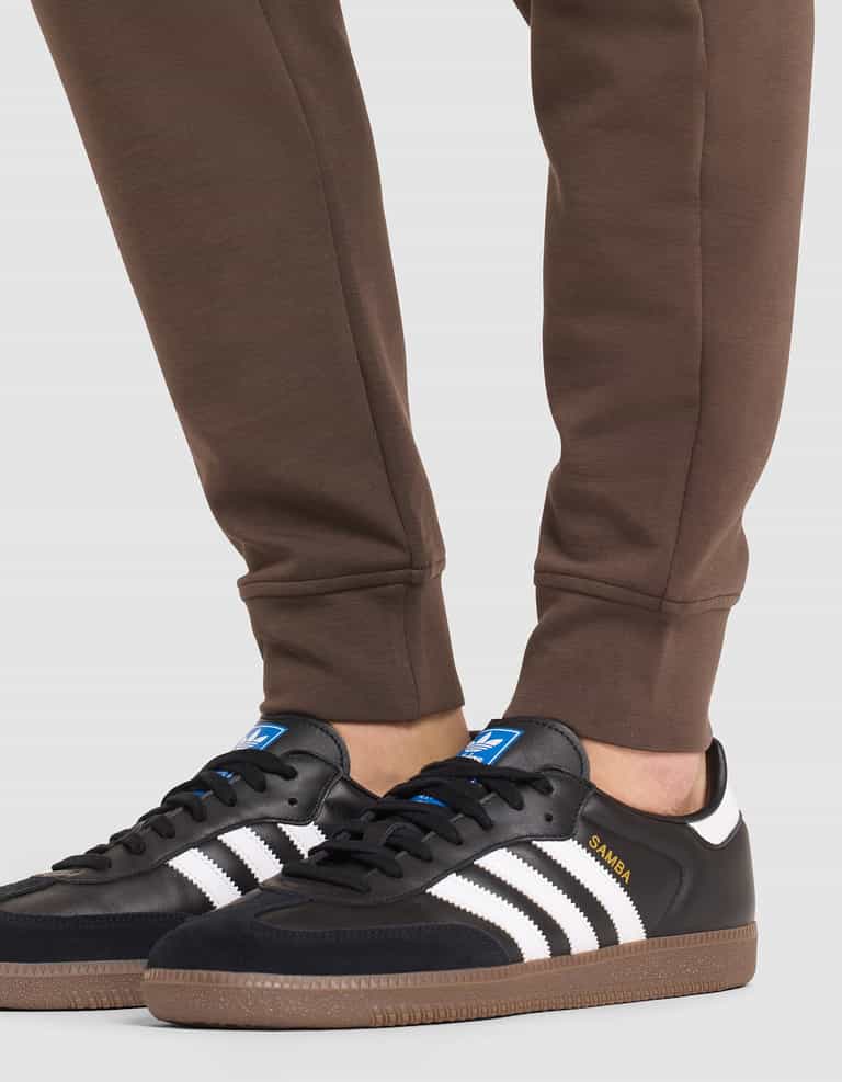 A person wearing Cozy Earth Men's StretchTech Jogger pants and black Adidas Samba sneakers with white stripes and gum soles stands on a light-colored surface. |Color:Cacao