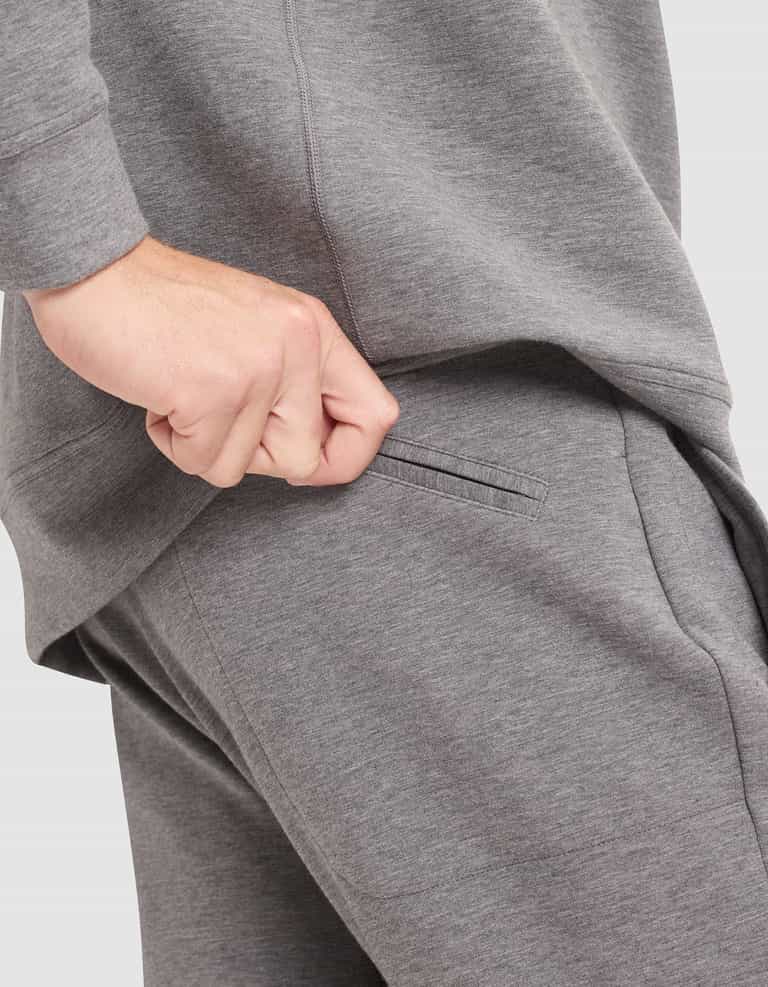 A person wearing Cozy Earth's Men's StretchTech Jogger in grey pulls at the waistband near the pocket, with their hand partially inside the pocket. |Color:Coal Heather