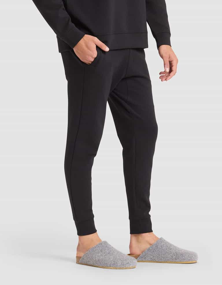 A person wearing Cozy Earth Men's StretchTech Jogger pants and a matching long-sleeve top stands with one hand in their pocket, wearing gray felt slippers with tan soles, on a white background. |Color:Jet Black