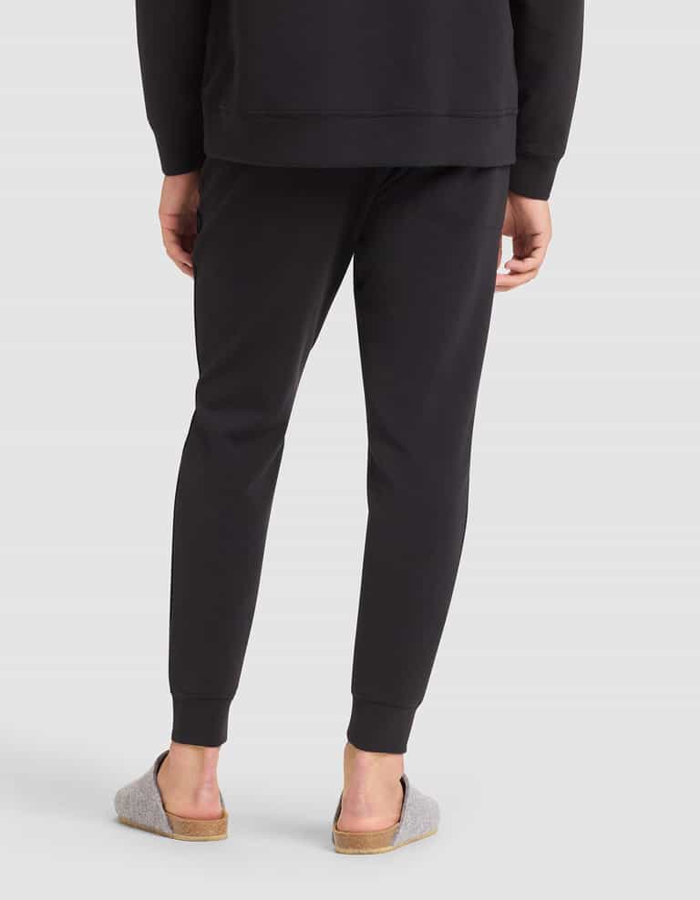 A person stands with their back to the camera, wearing Cozy Earth Men's StretchTech Jogger pants and a matching black sweatshirt, paired with light gray slippers with cork soles. The background is plain light gray. |Color:Jet Black