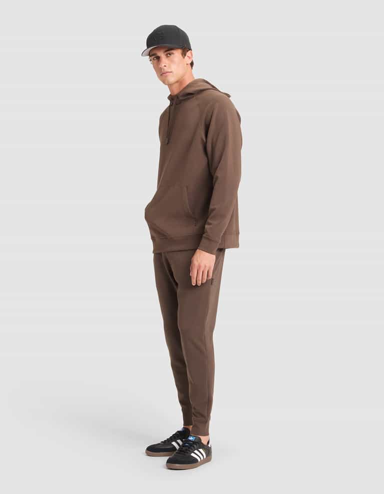 A man models the Cozy Earth Men's StretchTech Hoodie & Jogger Set in brown against a plain white background, paired with a black cap and black Adidas sneakers with white stripes. He stands with hands by his sides, gazing slightly to the side. |Color:Cacao
