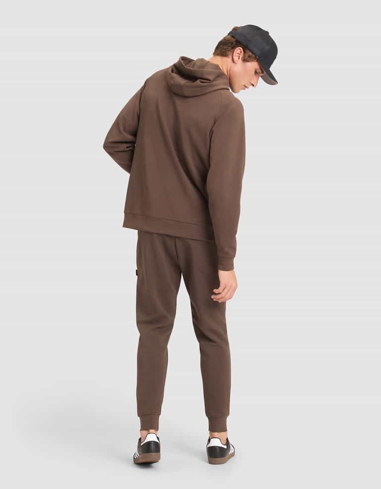 A man models the Cozy Earth Men's StretchTech Hoodie & Jogger Set in brown, paired with a black cap and black-and-white sneakers, standing against a plain light background and glancing over his shoulder. |Color:Cacao