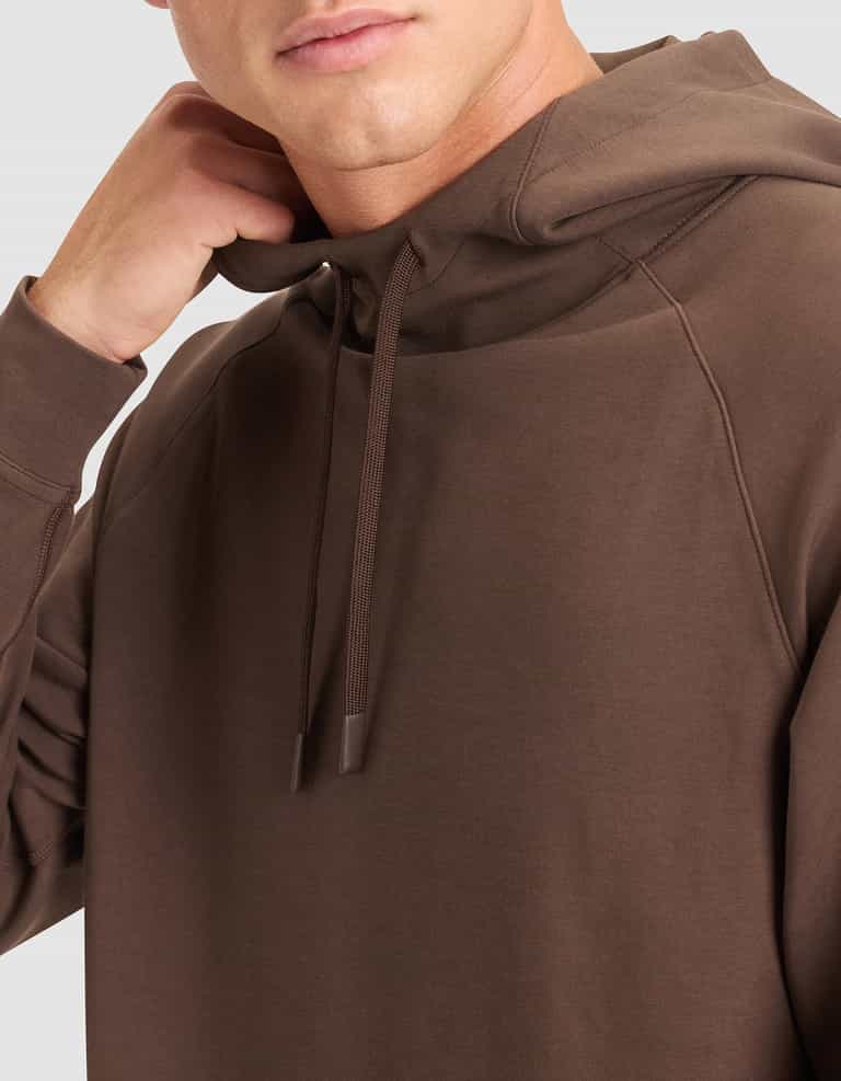 A person, shown from the shoulders up, touches their neck while wearing the Cozy Earth Men's StretchTech Hoodie with drawstrings. The background is light gray. |Color:Cacao
