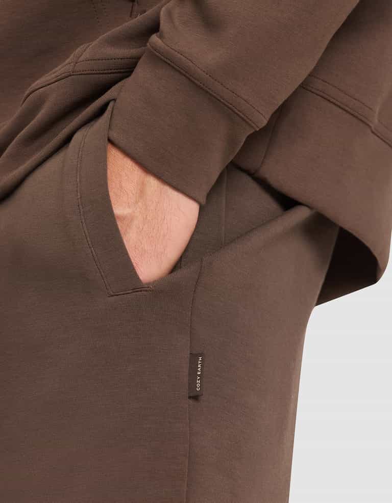 A person wears the Cozy Earth Men's StretchTech Hoodie & Jogger Set in brown, with one hand relaxed in the jogger pocket; a small tag on the pocket displays “COZY EARTH”. |Color:Cacao