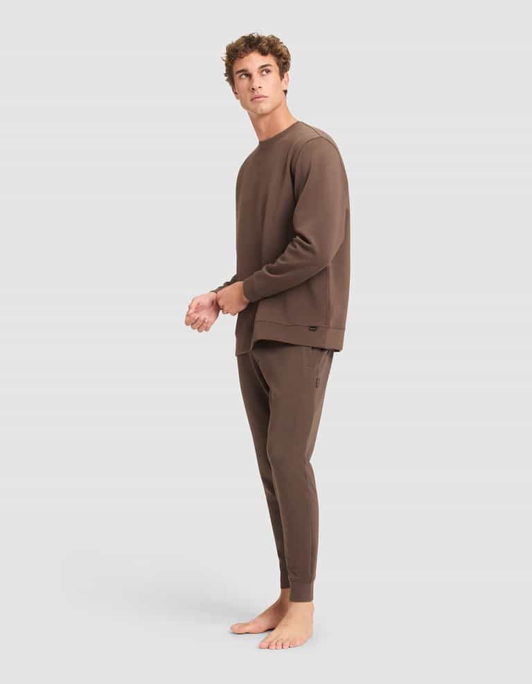 A person stands barefoot on a plain background, wearing the Cozy Earth Men's StretchTech Crewneck & Jogger Set in brown, looking slightly upward and to the side. |Color:Cacao