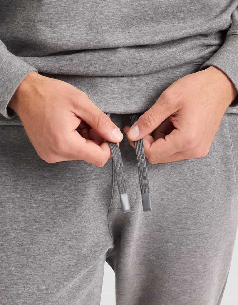 A man wearing the Cozy Earth Men's StretchTech Crewneck & Jogger Set in gray holds the jogger drawstrings, preparing to tie them. |Color:Coal Heather