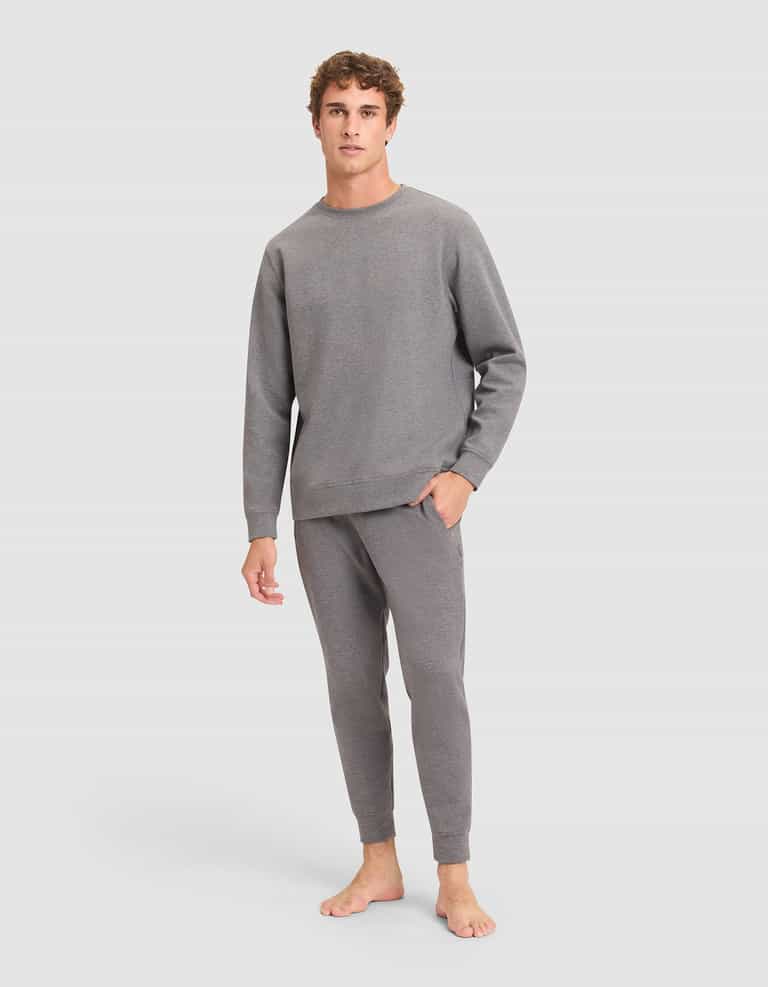 A young man with curly hair stands barefoot against a plain background, wearing the Cozy Earth Men's StretchTech Crewneck & Jogger Set in gray. |Color:Coal Heather