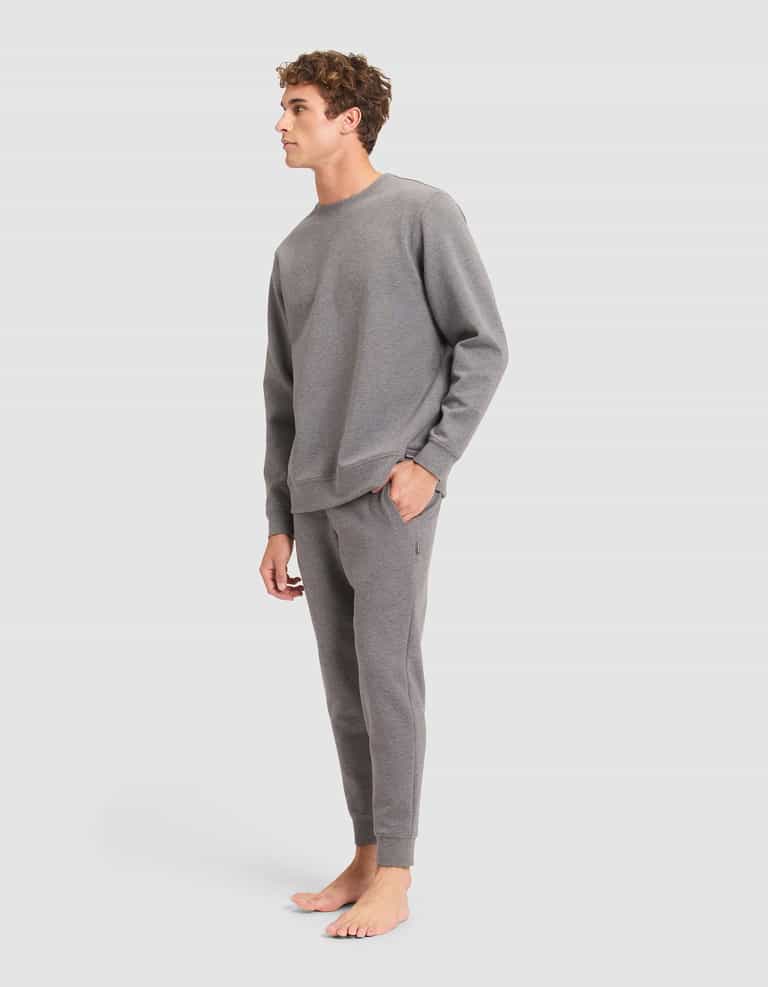 A person stands barefoot against a plain background, wearing the Cozy Earth Men's StretchTech Crewneck & Jogger Set in grey, with one hand in their pocket and looking to the side. |Color:Coal Heather
