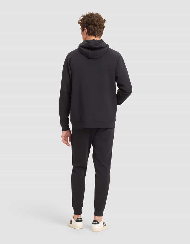 A man with curly hair faces away, wearing Cozy Earth's Men's StretchTech Hoodie & Jogger Set in black. He has one hand in his pocket and pairs the outfit with white sneakers featuring black accents against a plain light gray background. |Color:Jet Black