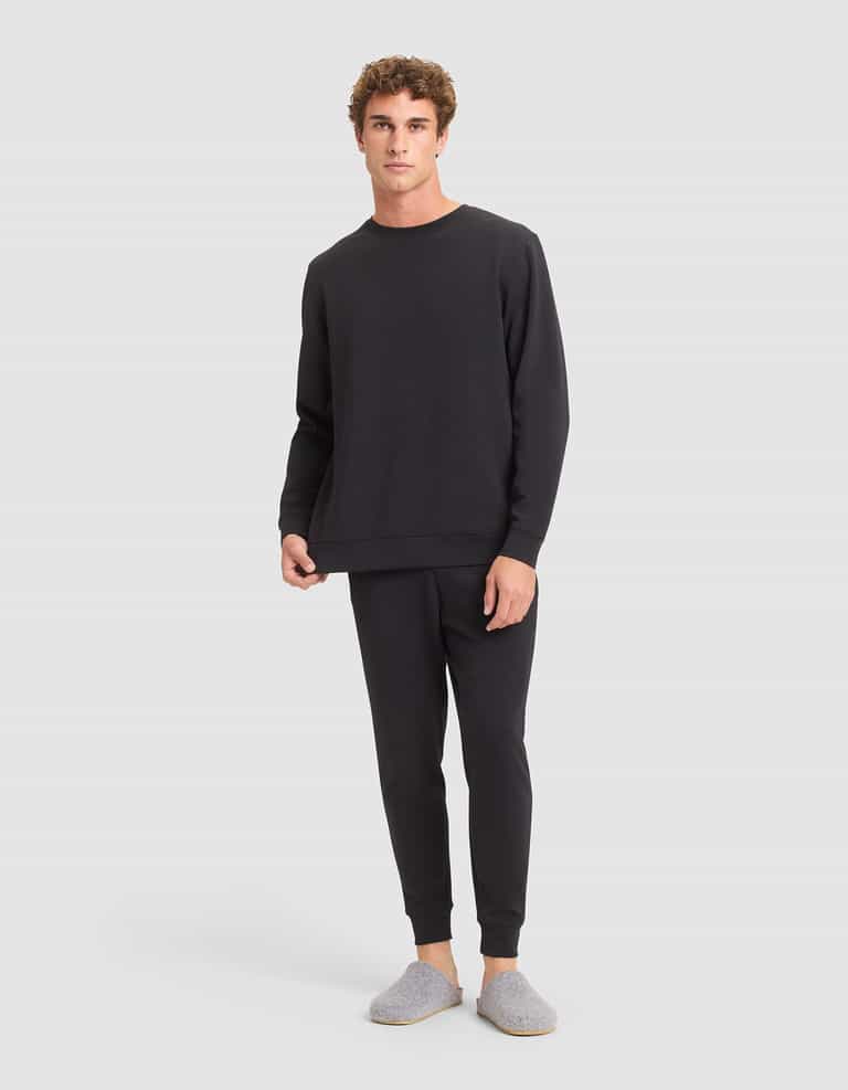 A man faces forward in a relaxed pose, wearing the Cozy Earth Men's StretchTech Crewneck & Jogger Set in black along with gray slippers, set against a plain background. |Color:Jet Black