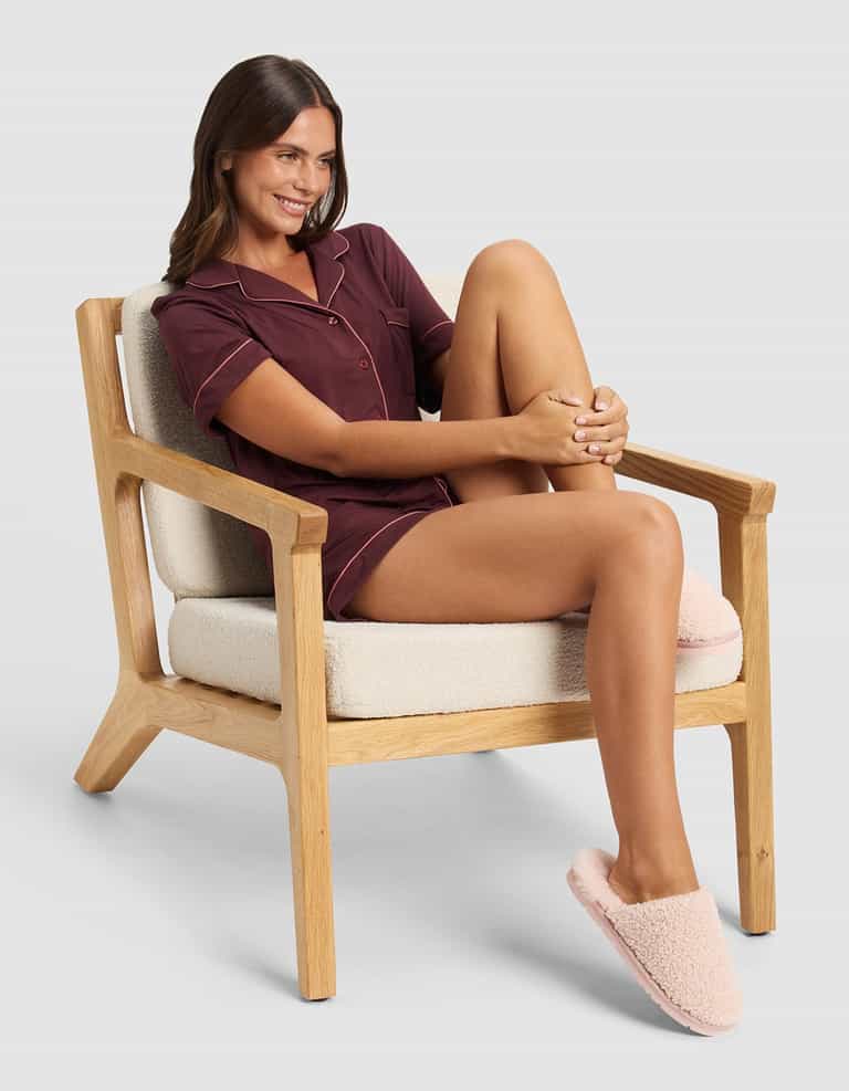 A woman wearing Cozy Earth's Puffy Sheep Slippers sits comfortably on a light cushioned wooden chair, smiling with one knee up. She is dressed in burgundy pajamas against a plain, light-colored background.|Color:Blossom