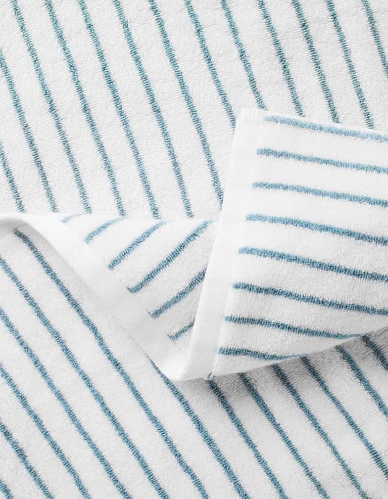 A close-up of Cozy Earth's Striped Luxe Bath Sheets in white with thin blue diagonal stripes, featuring a folded corner to highlight the textured detail. |Color:Brook Stripe