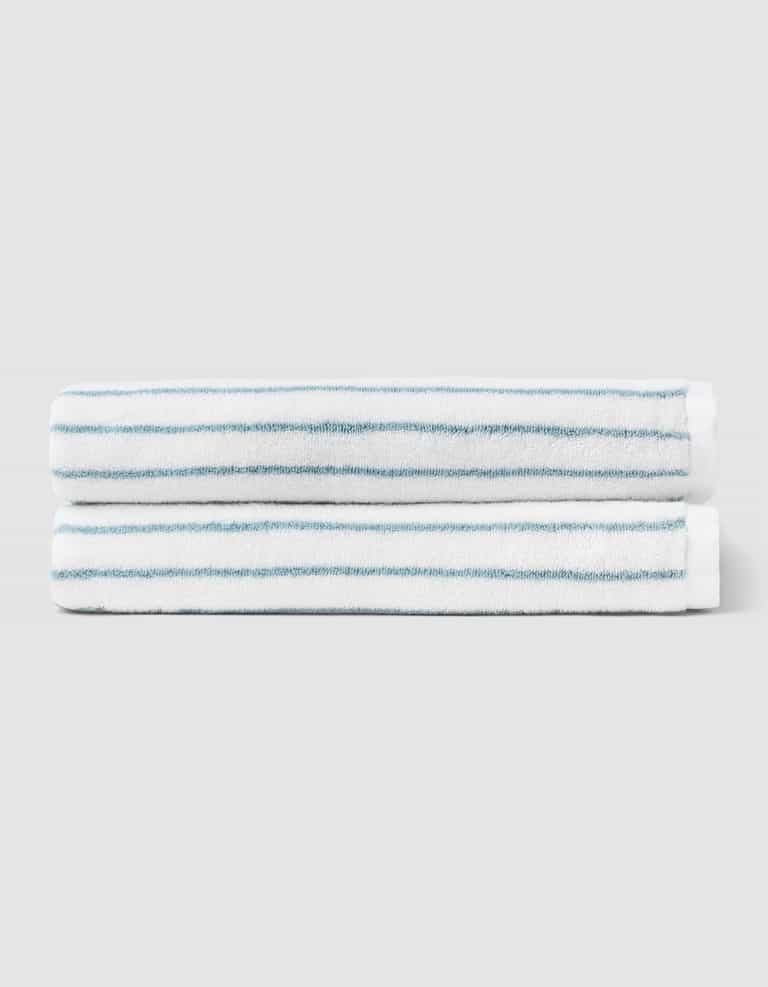 Two Striped Luxe Bath Towels by Cozy Earth, featuring thin blue stripes on white, are stacked together against a plain light gray background. |Color:Brook Stripe