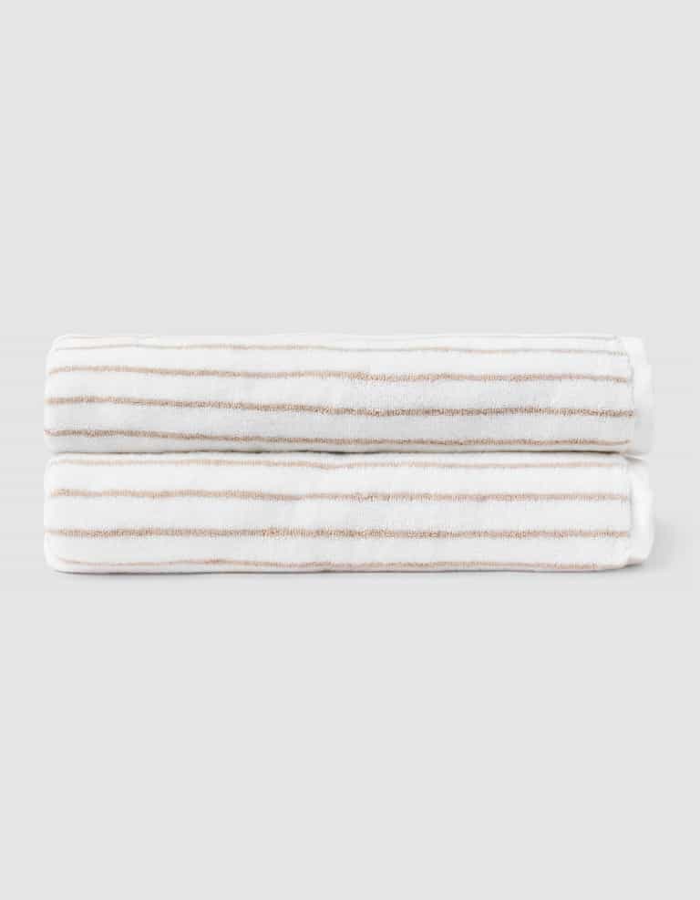Two Striped Luxe Bath Sheets by Cozy Earth, featuring white fabric with thin, evenly spaced tan stripes, are neatly folded and stacked against a plain light gray background. |Color:Sand Stripe