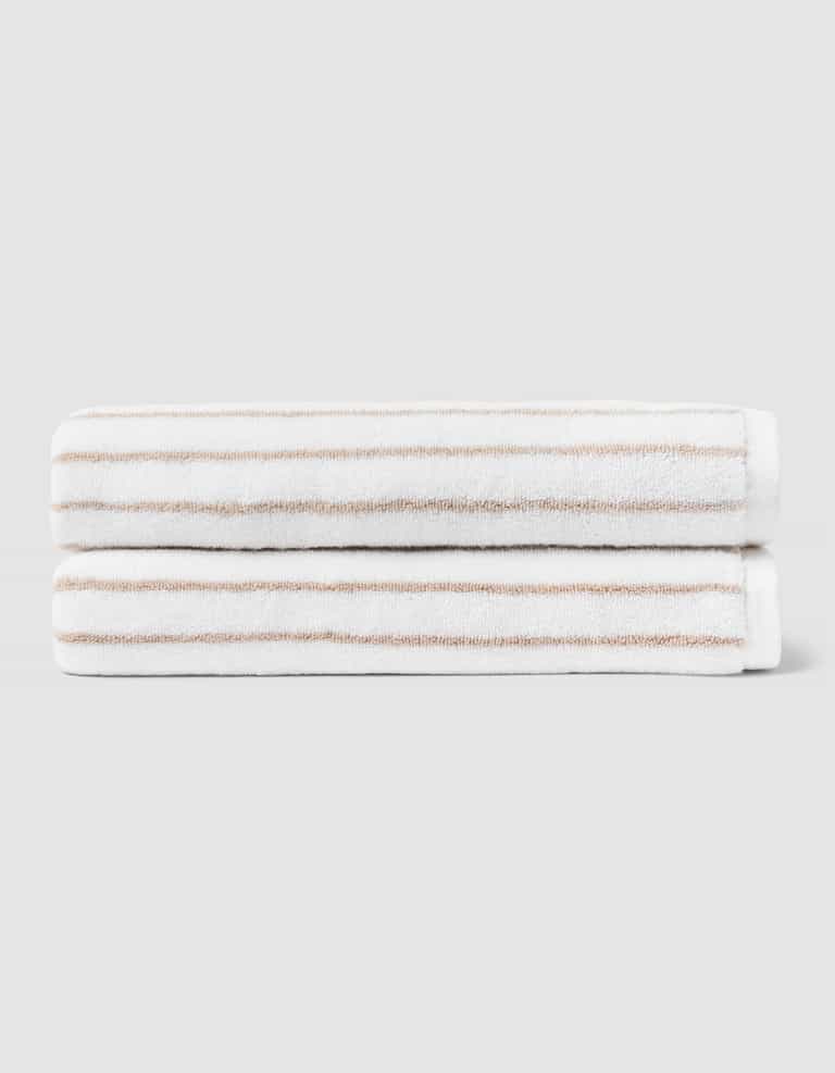 Two Striped Luxe Bath Towels by Cozy Earth, featuring thin light brown stripes on white, are stacked neatly against a plain light gray background. |Color:Sand Stripe