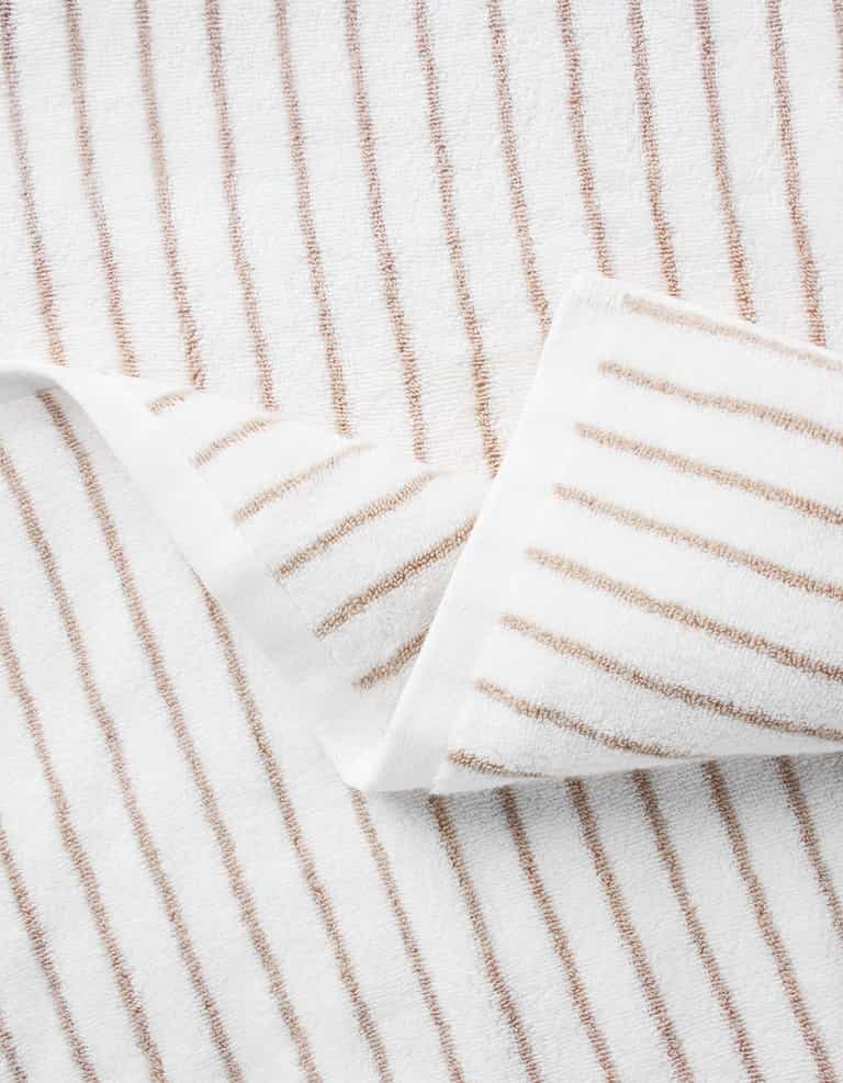 A close-up of the Cozy Earth Striped Luxe Bath Towel, featuring thin, evenly spaced brown vertical stripes on white fabric and a partial fold at one corner to display the pattern on both sides. |Color:Sand Stripe