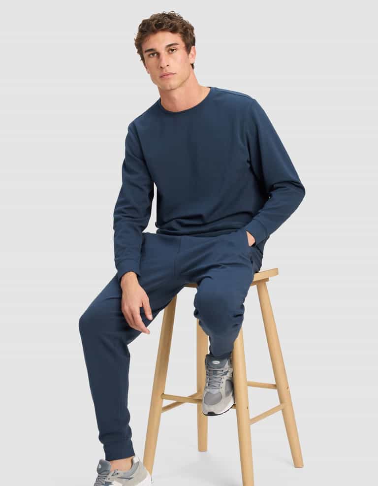 A young man wears the Cozy Earth Men's StretchTech Crewneck & Jogger Set in navy blue, paired with gray sneakers, while seated on a wooden stool against a plain light gray background, looking at the camera with one hand in his pocket.|Color:Eclipse