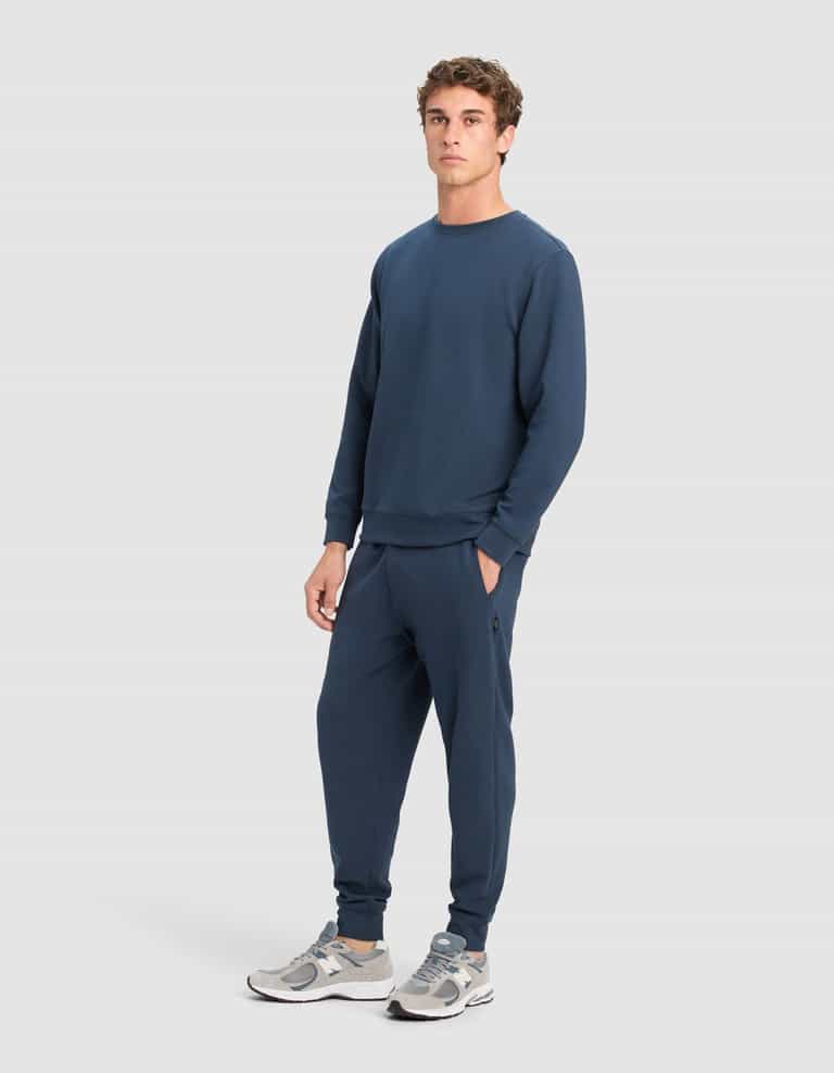 A man models the Cozy Earth Men's StretchTech Crewneck & Jogger Set in navy blue, paired with gray and white sneakers, standing facing forward with one hand in his pocket against a light gray background.|Color:Eclipse