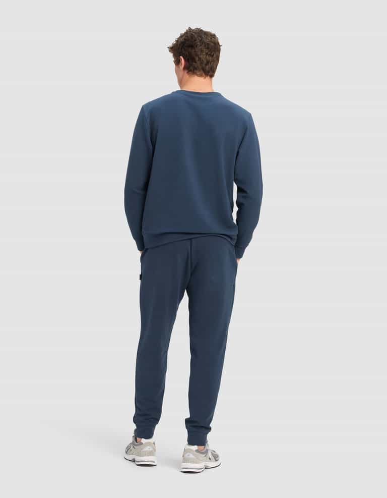 A man faces away, wearing Cozy Earth's Men's StretchTech Crewneck & Jogger Set in navy blue with white sneakers, set against a plain light gray background.|Color:Eclipse