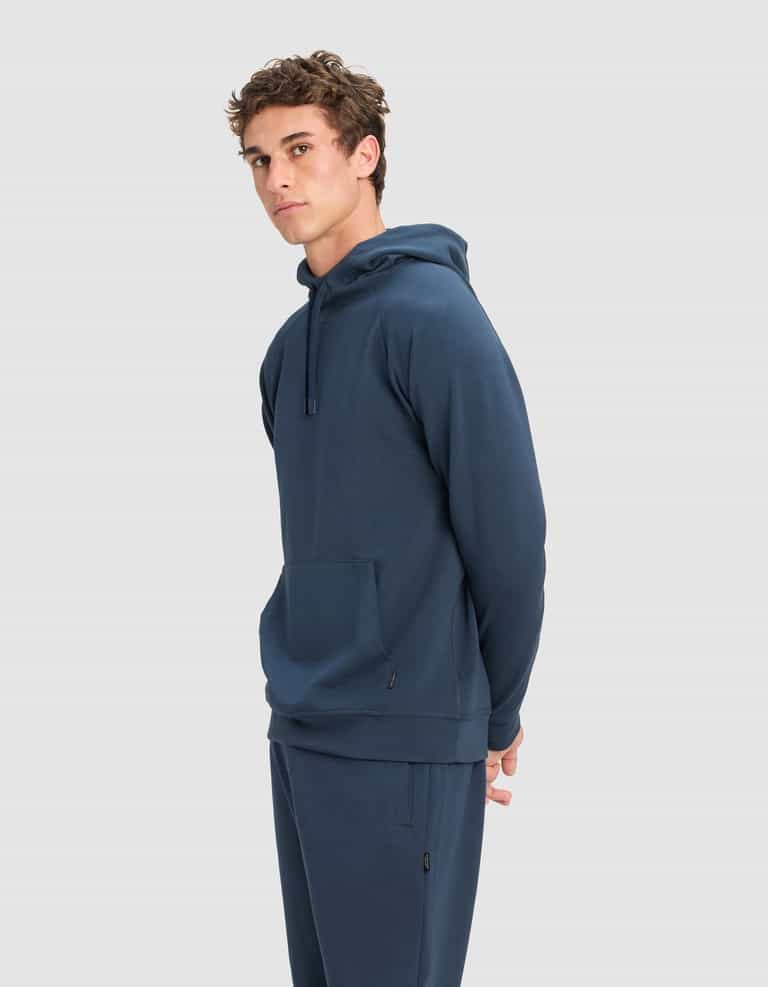 A young man with short, curly brown hair models the Cozy Earth Men's StretchTech Hoodie in blue, paired with matching pants, standing sideways with his hands behind his back against a light gray background.|Color:Eclipse
